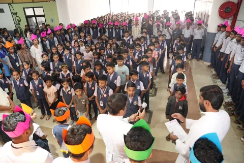 Young children in navy vests standing in rows facing two men in white, one holding a microphone