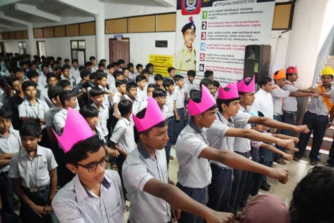 Students in school uniform, several in pink paper crowns, standing with their arms stretched forward in front of a wall poster