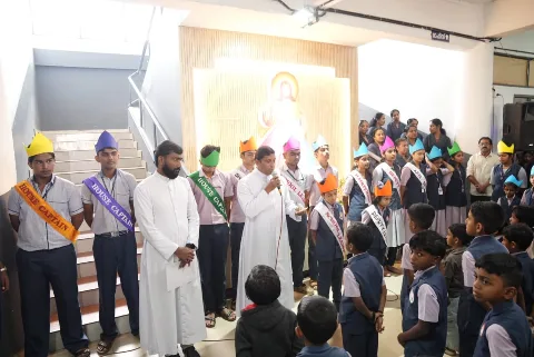Students in orange and purple House Captain sashes and paper crowns beside two men in white