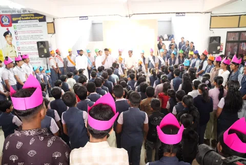 Students standing in rows facing a line of students in paper crowns and sashes, with a man in white speaking into a microphone