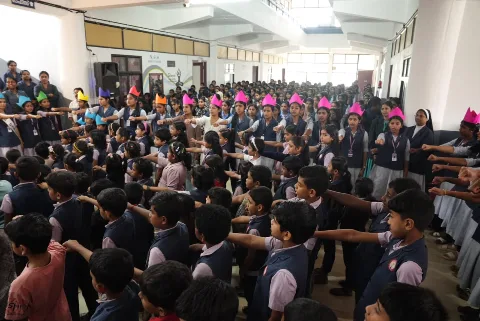 Girls in navy uniform, some in pink paper crowns, standing with their arms stretched forward in a hall with white pillars